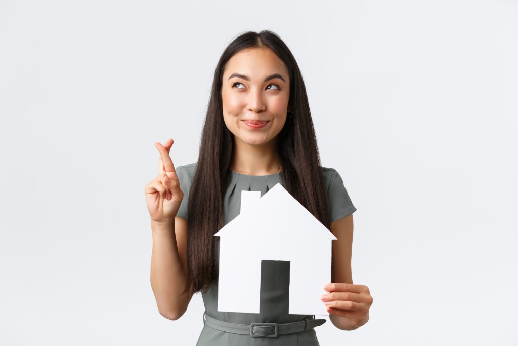 Insurance, loan, real estate and family concept. Hopeful and optimistic smiling asian woman cross fingers and smiling, dreaming of finding new home, holding paper house, making wish